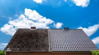 Toiture en tuiles sombres à moitié démoussée sous un ciel bleu